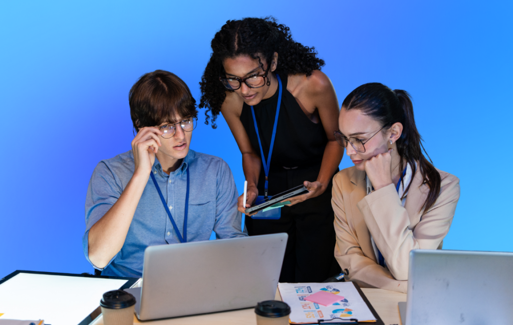 Three colleagues working together at a desk with laptops and documents against a blue background.