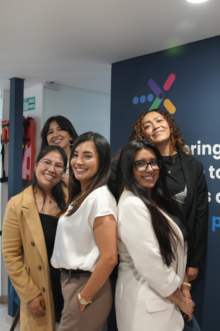 Five smiling women standing together in a modern office space with a branded wall.