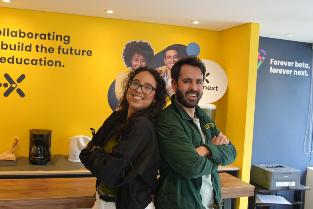 Two colleagues standing back-to-back and smiling in a modern office with yellow walls featuring company branding.