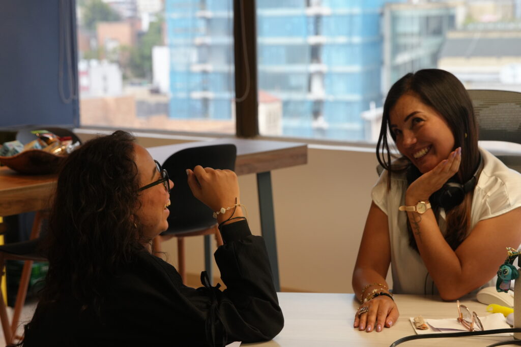 Two colleagues smiling and talking while sitting at a desk in a bright office with large windows.