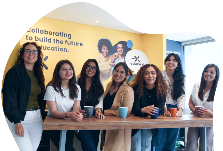 Seven team members standing together at a wooden table in an office with a yellow wall featuring the edunext logo.