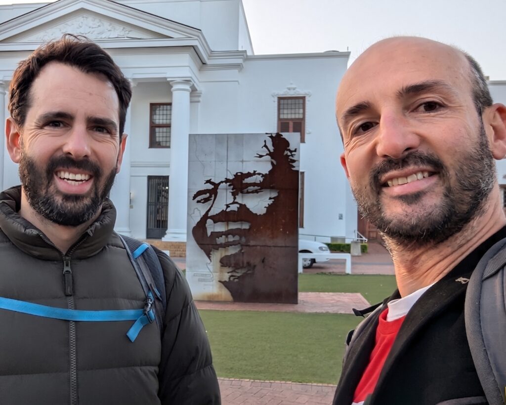 Two men smiling in front of a white building and a metal sculpture.