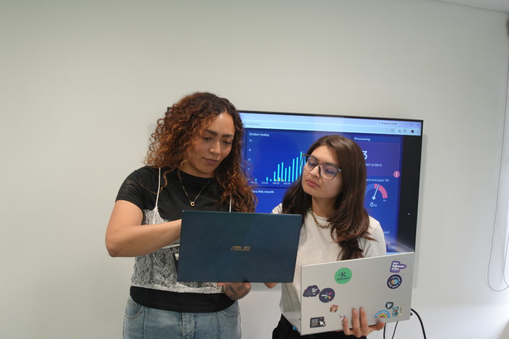 Two women standing in an office, one holding an ASUS laptop and the other holding a laptop with edunext stickers, while looking at a large monitor displaying data analytics.