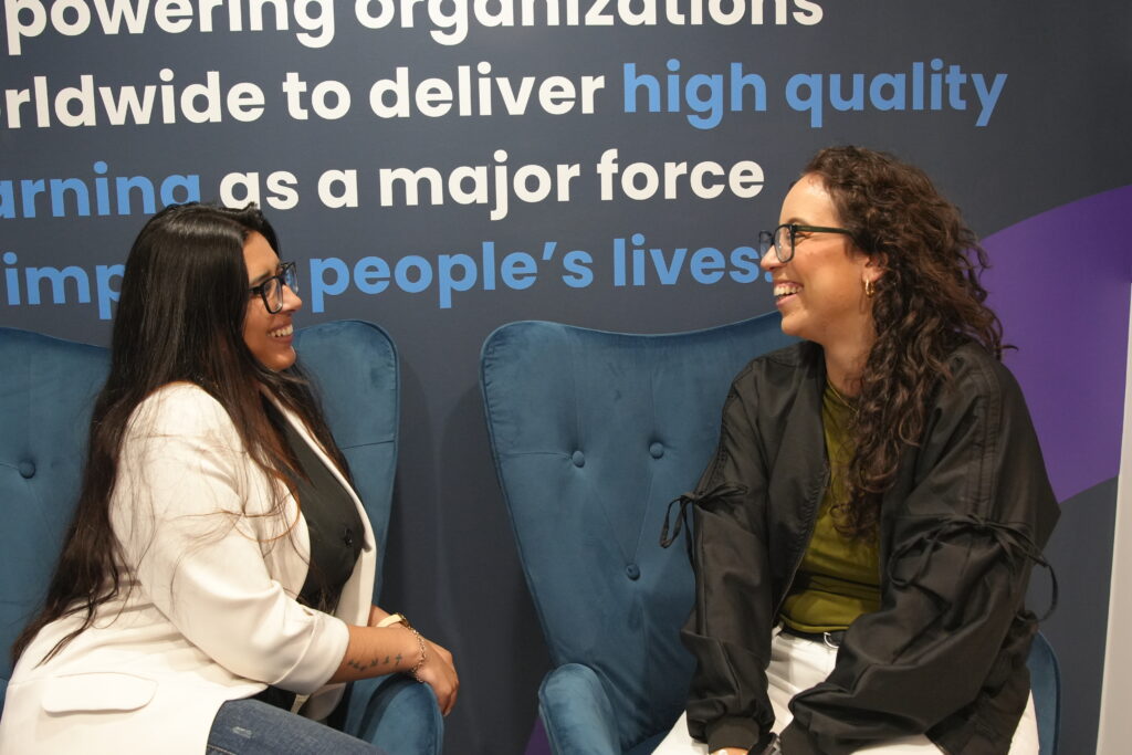 Two women sitting in blue armchairs and talking in front of a branded wall.