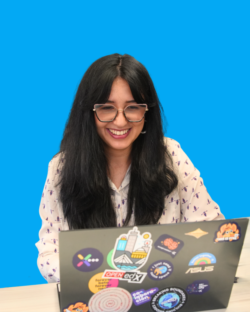 A person with long dark hair and glasses smiling while working on a laptop covered in various stickers, including Open edX branding.
